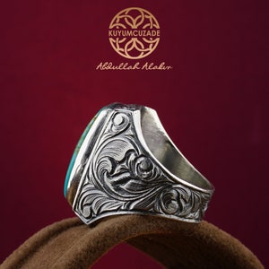 May include: A silver ring with a turquoise stone set in a detailed, engraved design. The ring is on a brown surface.