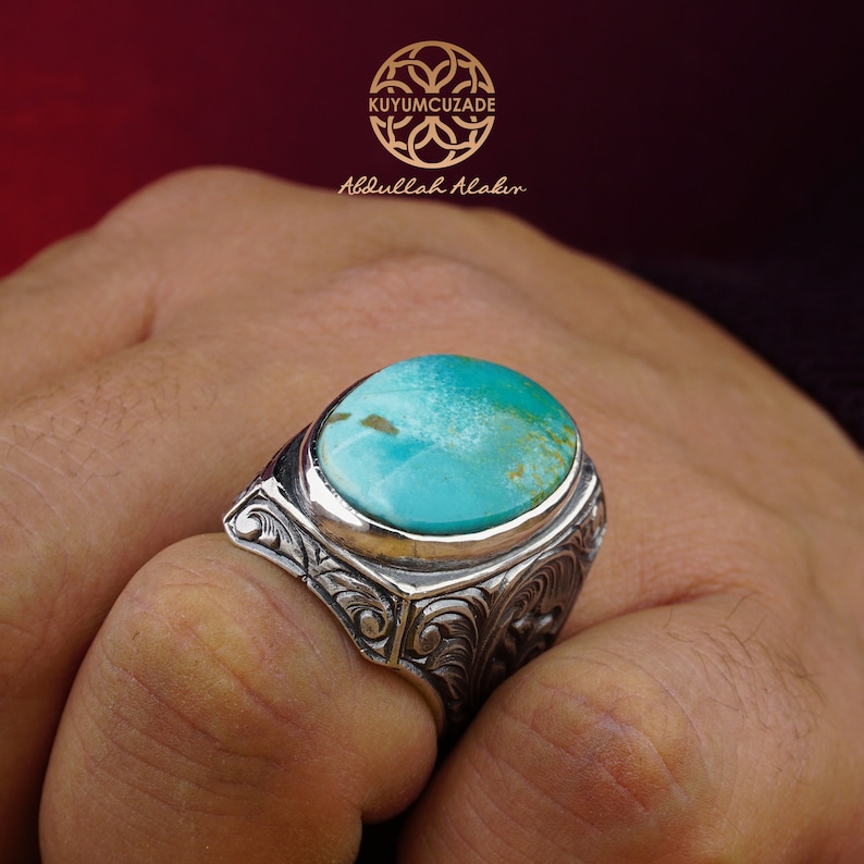 May include: A silver ring with a large turquoise gemstone set in a detailed, ornate design. The ring is on a person's finger.