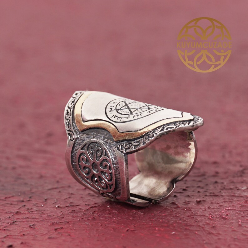 May include: A silver ring with a gold-plated shield design featuring intricate details and Hebrew lettering. The ring has a wide band with a decorative pattern.
