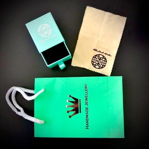 May include: A turquoise gift bag with a white handle and a crown logo that says "Handmade Jewellery". The bag is next to a turquoise box with a black interior and a silver logo that says "Kuyumcuzade Abdullah Akar". There is also a beige polishing cloth with a silver logo that says "Kuyumcuzade Abdullah Akar".