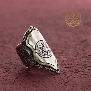 May include: A silver ring with a gold-plated shield featuring a Star of David and Hebrew text. The ring has intricate details and a wide band.