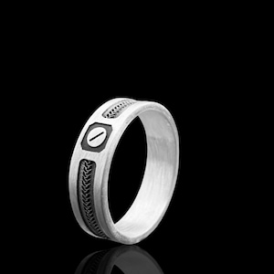 Könnte beinhalten: Ein silberner Ring mit detailliertem Design. Der Ring hat ein zentrales rechteckiges Element mit einer horizontalen Linie, flankiert von dekorativen Mustern. Das Band hat ein strukturiertes, gewebtes Muster. Der Ring ist vor einem schwarzen Hintergrund platziert.