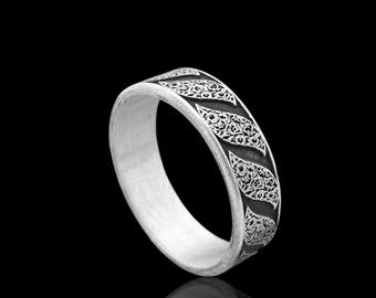May include: A silver ring with a detailed, repeating pattern of black filigree. The ring is set against a black background, with a reflection visible below. The band has a flat profile.
