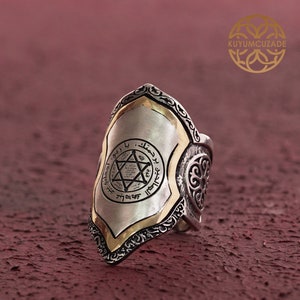 May include: A silver ring with a shield-shaped design. The shield has a gold border and a six-pointed star with Arabic script inside. The ring has intricate details and a textured finish.