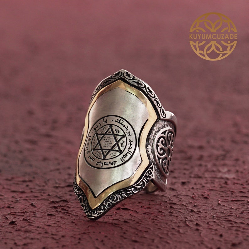 May include: A silver ring with a shield-shaped design. The shield has a gold border and a six-pointed star with Arabic script inside. The ring has intricate details and a textured finish.