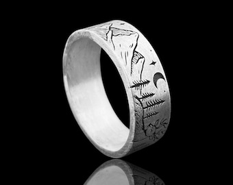 May include: A silver ring with a mountain and forest design. The ring features a detailed engraving of a mountain range, trees, a crescent moon, and stars. The ring is approximately 0.3 inches wide.