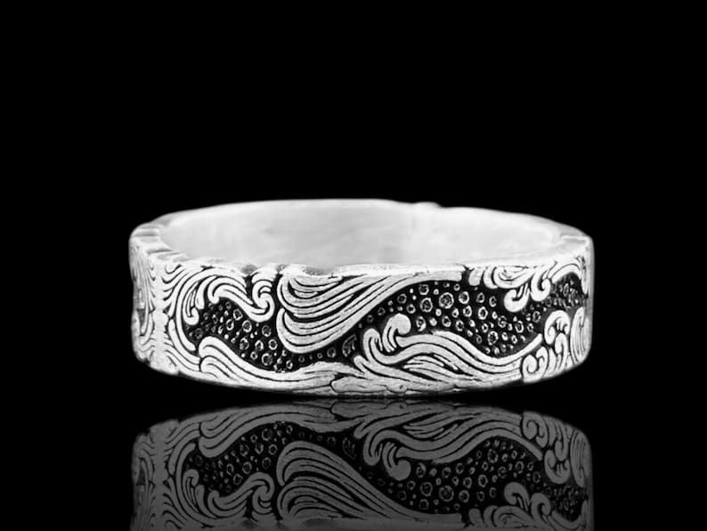 May include: A silver ring with an engraved wave pattern. The ring's design features flowing lines and dotted details, creating a textured appearance. The ring is displayed against a black background, with its reflection visible.