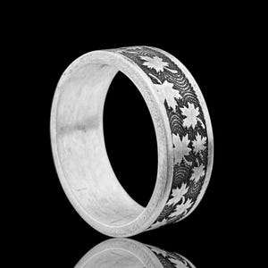Puede incluir: Un anillo de plata con un patrón detallado de hojas de arce y líneas onduladas. El anillo es grueso y tiene un acabado pulido. El diseño está grabado en la banda, creando un contraste.