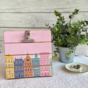 May include: A pink wooden clipboard with a silver clip and a painted design of colorful houses. The houses are yellow, blue, orange, teal, and pink.