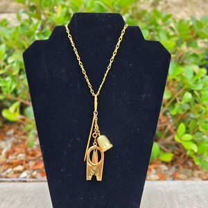 Könnte beinhalten: Eine goldfarbene Halskette mit einem Anhänger mit Näh-Themen-Anhängern. Der Anhänger enthält eine Schere, einen Fingerhut und eine Nadel. Die Halskette wird auf einem schwarzen Samtständer vor einem Hintergrund aus grünem Laubwerk präsentiert.