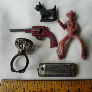 May include: A collection of vintage toys including a black dog figurine, a red toy revolver, a pink cowboy figure, a metal ring with a clear stone, and a silver-toned harmonica marked PeeWee Made in Occupied Japan, next to a wooden ruler.