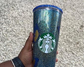 May include: A blue and gold glitter tumbler with a Starbucks logo and butterfly designs. The tumbler has a blue rim with rhinestones.