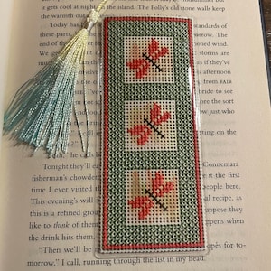 May include: A handmade bookmark with a clear plastic cover. The bookmark features three embroidered squares with red dragonfly designs on a green background. A tassel hangs from the top. The bookmark is resting on the pages of a book.