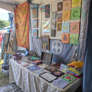 May include: A vendor booth displays various art pieces, including colorful square paintings with whimsical animal illustrations. A table is covered with artwork, including prints and small framed pieces. The booth is set up under a tent with decorative textiles.