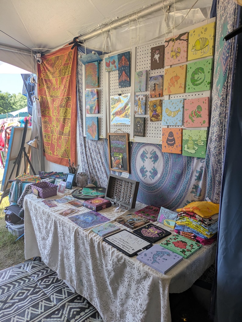 May include: A vendor booth displays various art pieces, including colorful square paintings with whimsical animal illustrations. A table is covered with artwork, including prints and small framed pieces. The booth is set up under a tent with decorative textiles.