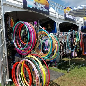 May include: A display of colorful hula hoops in various sizes and designs. The hoops are arranged on metal racks and are a mix of solid colors and patterns. Clothing items hang on a rack in the background, with signs that read "Mind Hydration" and "Stone Goods".