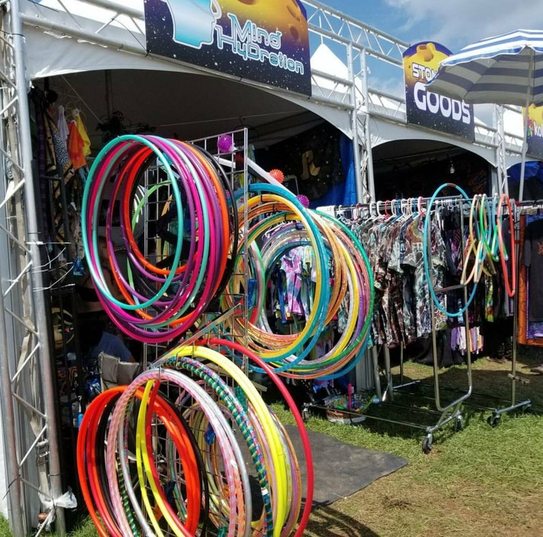 May include: A display of colorful hula hoops in various sizes and designs. The hoops are arranged on metal racks and are a mix of solid colors and patterns. Clothing items hang on a rack in the background, with signs that read "Mind Hydration" and "Stone Goods".