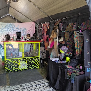May include: A vendor booth at an outdoor market, displaying a variety of colorful clothing and accessories. The booth features a counter with the text "Vibrant Hops" and a display of hats, scarves, and other items. The tent has a patterned rug.