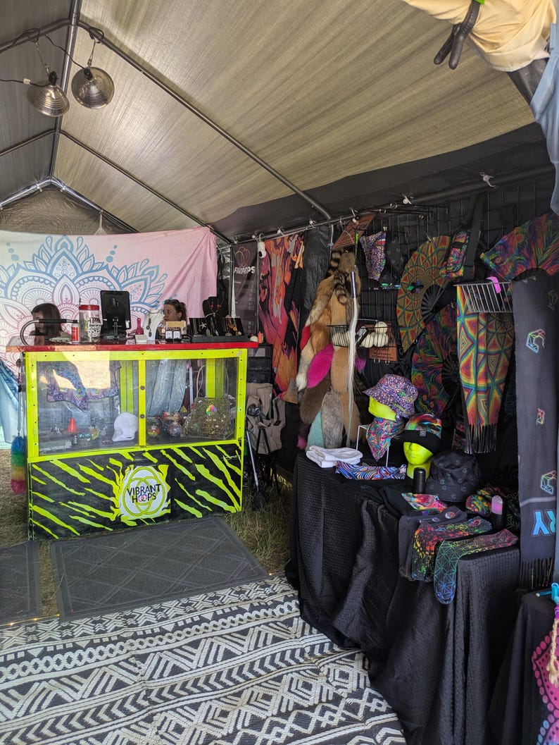 May include: A vendor booth at an outdoor market, displaying a variety of colorful clothing and accessories. The booth features a counter with the text "Vibrant Hops" and a display of hats, scarves, and other items. The tent has a patterned rug.