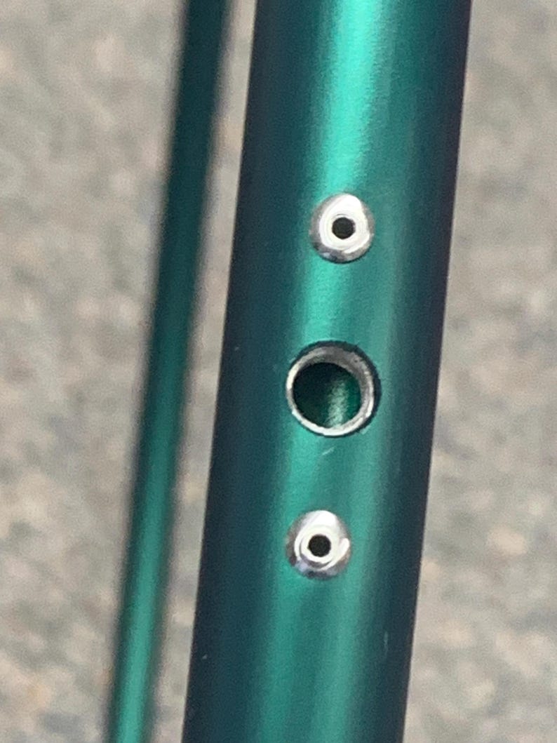 Puede incluir: Primer plano de un tubo met&aacute;lico verde con tres ojales plateados. Los ojales est&aacute;n espaciados uniformemente a lo largo del tubo.