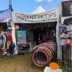 May include: A vendor booth with a white tent displaying colorful hula hoops and tie-dye apparel. The sign reads "VIBRANT HOOPS" and "Zero-Waste Juggle & Flow Props." A cooler sits in front of the booth. The sky is blue.