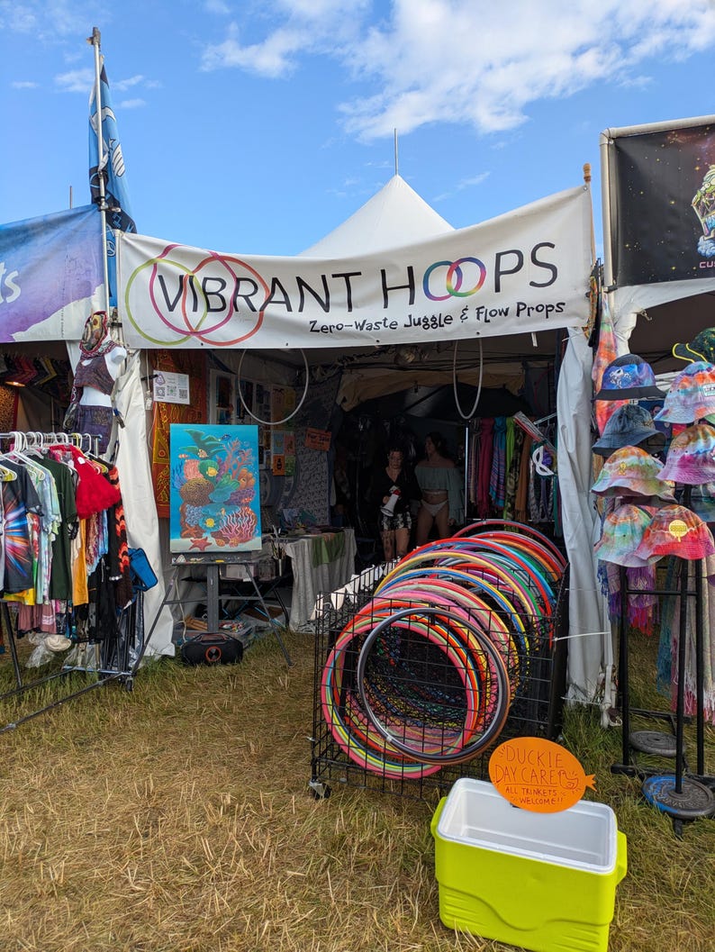 May include: A vendor booth with a white tent displaying colorful hula hoops and tie-dye apparel. The sign reads "VIBRANT HOOPS" and "Zero-Waste Juggle & Flow Props." A cooler sits in front of the booth. The sky is blue.