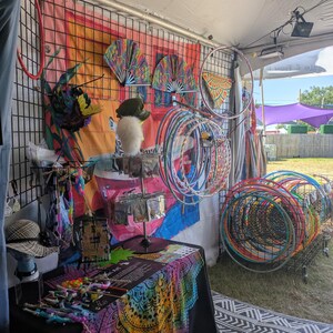 May include: A vendor booth displays colorful hula hoops, fans, and accessories. The display includes a variety of hoops in different sizes and colors, along with decorative fans and other items. A table is covered with a vibrant, patterned cloth.