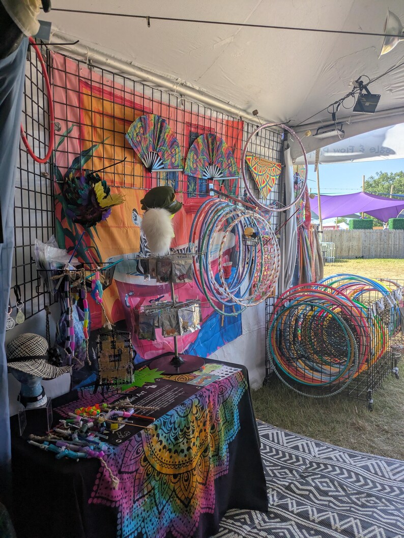 May include: A vendor booth displays colorful hula hoops, fans, and accessories. The display includes a variety of hoops in different sizes and colors, along with decorative fans and other items. A table is covered with a vibrant, patterned cloth.