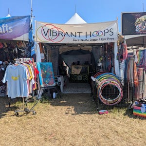 May include: A vendor booth with a banner that reads "VIBRANT HOOPS Zero-Waste Juggle & Flow Props." The booth displays colorful hula hoops, clothing, and art. The scene is set outdoors on a sunny day.