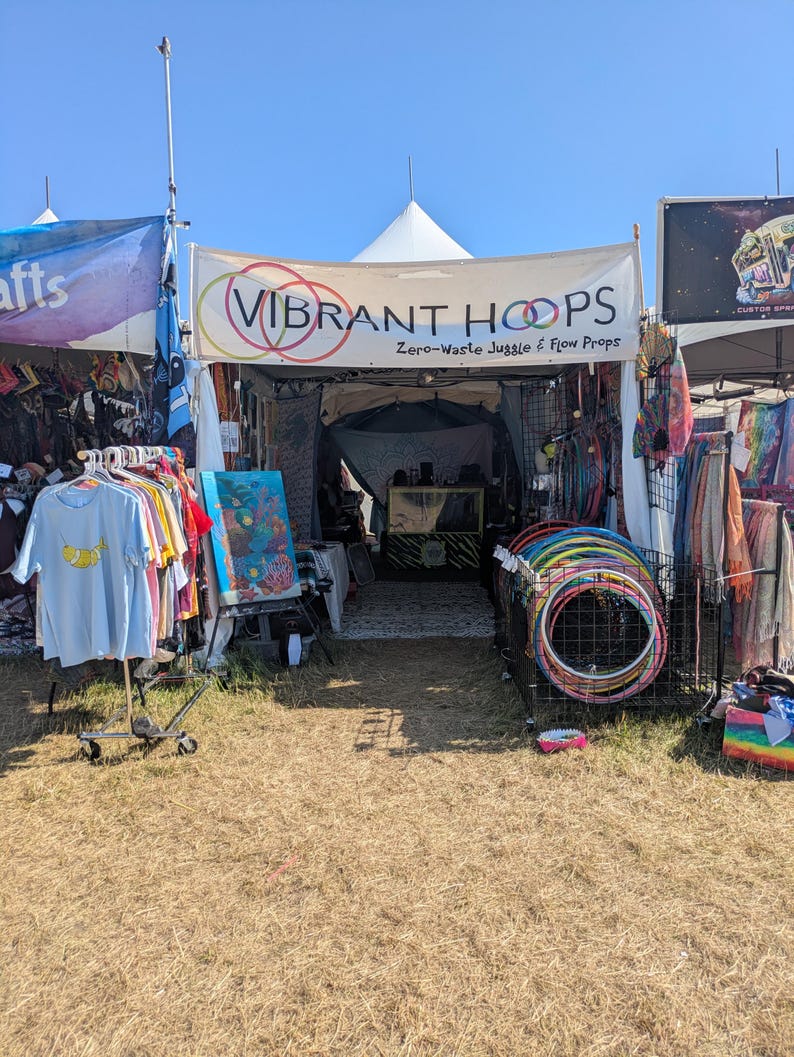 May include: A vendor booth with a banner that reads "VIBRANT HOOPS Zero-Waste Juggle & Flow Props." The booth displays colorful hula hoops, clothing, and art. The scene is set outdoors on a sunny day.