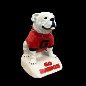 May include: A white bulldog statue wearing a red jersey with a black "G" and silver spiked collar. The statue sits on a white base with the words "GO DAWGS" in red. The bulldog has a black nose and a pink tongue.