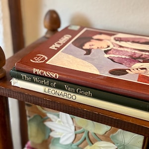 Puede incluir: Una pila de tres libros de arte de tapa dura. El libro superior es marrón con el texto PICASSO y una ilustración. El libro del medio es verde oscuro con el texto The World of Van Gogh. El libro inferior es blanco con el texto LEONARDO.