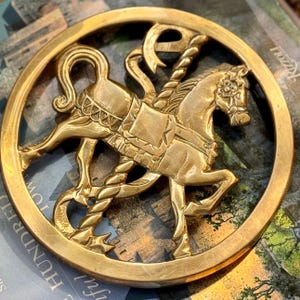 May include: A gold-colored, circular decorative piece featuring a carousel horse design. The horse is detailed with a saddle, bridle, and floral accents. The piece is set within a circular frame.
