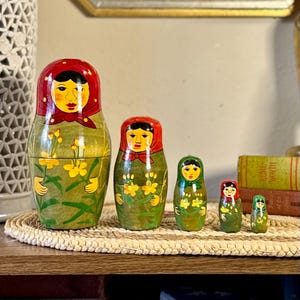 May include: A set of five nesting dolls, each painted with a traditional design. The largest doll is green with a red head covering and a yellow floral pattern. The dolls decrease in size, with similar colors and patterns.