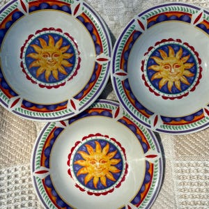 May include: Three round ceramic bowls featuring a central yellow sun face design on a blue background. The rims are decorated with a scalloped red border, a band of blue circles, and a green leaf pattern on a white base with red accents.