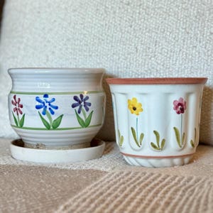 May include: Two ceramic flower pots. One is white with a green rim and painted flowers in red, blue, and purple. The other is white with a pink rim and painted yellow and pink flowers. Both have green stems.