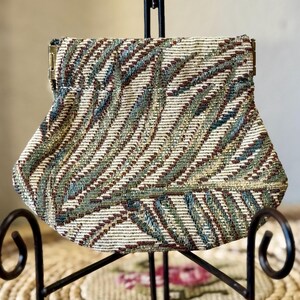 May include: A vintage clutch purse with a woven fabric design. The purse features a mix of colors, including cream, brown, and teal, arranged in a diagonal pattern. It has a metal clasp closure and a rounded bottom.