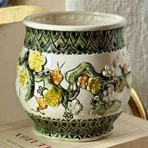 May include: A ceramic planter with a white base featuring raised, three-dimensional floral branches in yellow, orange, and white. The top and bottom rims are decorated with a dark green, intricate lattice pattern that encircles the entire vessel.