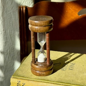 May include: A small, wooden hourglass with a clear glass center filled with white sand. The hourglass has a dark brown finish and is set on a vintage book. The design features a classic, time-telling aesthetic.