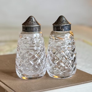 May include: A pair of clear glass salt and pepper shakers with a diamond-cut pattern. Each shaker has a silver-colored metal lid with small holes. The shakers are displayed on a brown surface, suggesting a vintage or antique aesthetic.