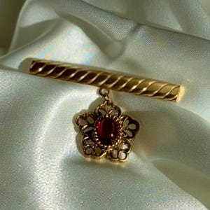 May include: A gold-toned bar pin with a floral pendant. The bar has a textured design, and the pendant features a red gemstone center surrounded by an openwork floral design. The pin is set against a soft, white background.
