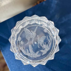 May include: A clear, faceted glass dish with an etched butterfly and floral design. The dish has a circular interior and a multi-sided, starburst-like rim. The butterfly design is surrounded by etched flowers and leaves.