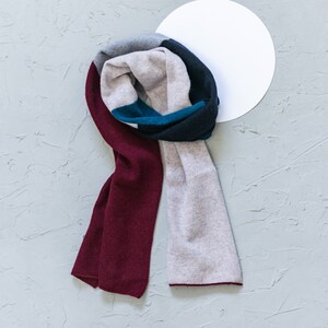 May include: A multi-colored scarf with burgundy, gray, black, and teal stripes. The scarf is folded and arranged on a gray textured surface.