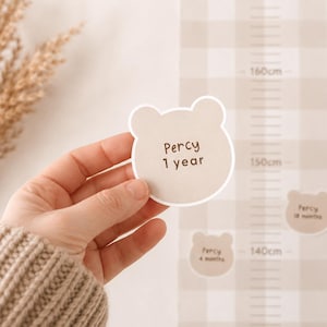 May include: A beige bear-shaped sticker with the text Percy 1 year, held in front of a beige and white gingham growth chart featuring metric measurements from 140cm to 160cm, with additional bear-shaped stickers labeled Percy 4 months and 18 months.