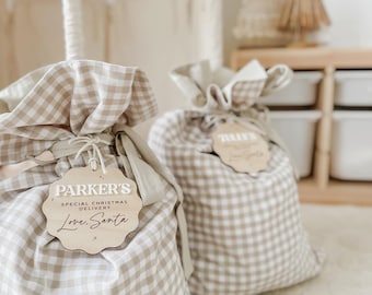 May include: Two beige and white gingham fabric gift bags with wooden tags. The tags read "Parker's Special Christmas Delivery Love, Santa". The bags are tied with a natural twine and are sitting on a cream-colored surface.