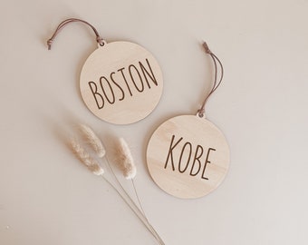 May include: Two round wooden ornaments with the names "BOSTON" and "KOBE" engraved in a simple font. Each ornament has a small hole at the top with a brown string for hanging. The background is a neutral beige.