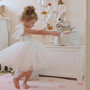 May include: A young child wearing a white tulle dress with bow accents, twirling on a pink rug with heart patterns. A white storage chest and a wall organizer with small gifts are in the background.