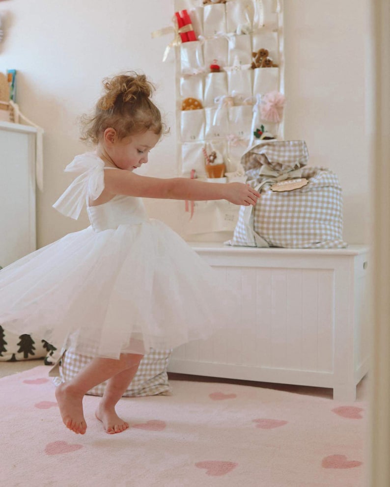 May include: A young child wearing a white tulle dress with bow accents, twirling on a pink rug with heart patterns. A white storage chest and a wall organizer with small gifts are in the background.