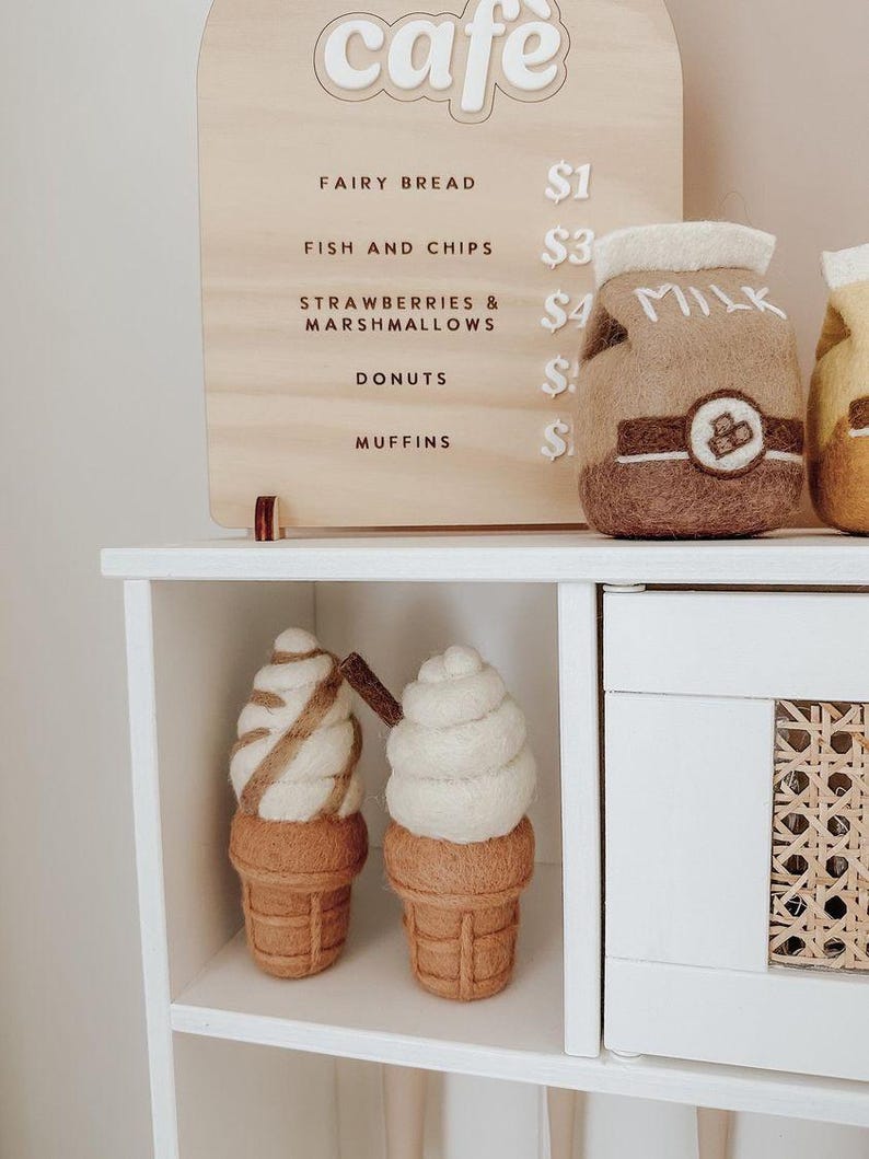 Pu&ograve; includere: Un cartello di legno bianco con la parola "cafe" in un cerchio bianco. Il cartello elenca i prezzi per il pane delle fate, il pesce e le patatine fritte, le fragole e i marshmallow, i donut e i muffin. Due coni gelato in feltro sono su uno scaffale bianco. Un cono gelato &egrave; bianco con vortici marroni e l'altro &egrave; marrone con vortici bianchi. Un cartone di latte in feltro &egrave; sullo scaffale dietro i coni gelato.