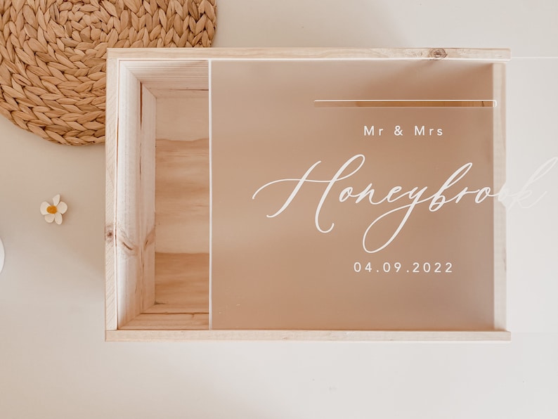Puede incluir: Una caja de madera con una tapa de acr&iacute;lico transparente. La tapa tiene el texto "Mr & Mrs Honeymoon 04.09.2022" escrito en blanco.
